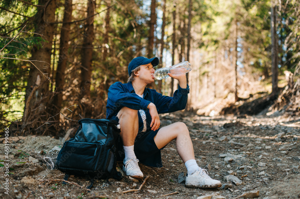 Handsome young male tourist relaxes sitting on a trail in the mountains with a backpack and quenches thirst by drinking water from a bottle and looks away.