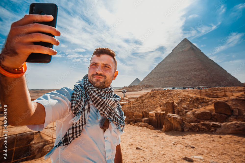 A young man, in front of the pyramid, raises his phone, takes a selfie with his fingers in front of the camera. - obrazy, fototapety, plakaty 