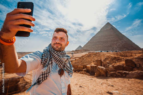 A young man, in front of the pyramid, raises his phone, takes a selfie with his fingers in front of the camera.
