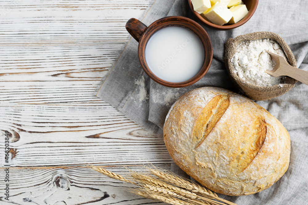 Fresh white bread with a crisp crust on a light napkin. A mug with milk, butter in a clay bowl and flour in a canvas bag in the background. Top view