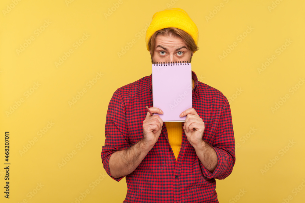 Portrait of hipster bearded guy in beanie hat and checkered shirt standing covering half of face with paper notebook, looking at camera. Indoor studio shot isolated on yellow background. Portrait of hipster bearded guy in beanie hat and checkered shirt standing covering half of face with paper notebook, looking at camera. Indoor studio shot isolated on yellow background.