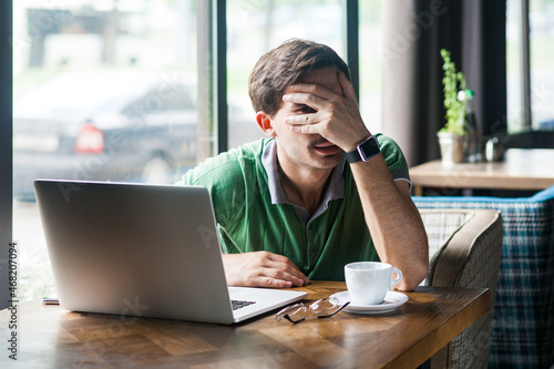 Portrait of upset businessman wearing green T-shirt, covering face, looking through fingers with big eyes, expressing sorrow and sadness. Indoor shot near big window, cafe background.