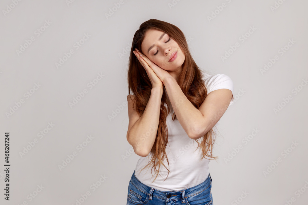Portrait of beautiful woman sleeping laying down on her palms, having comfortable nap and resting, dozing off, wearing white T-shirt. Indoor studio shot isolated on gray background. Portrait of beautiful woman sleeping laying down on her palms, having comfortable nap and resting, dozing off, wearing white T-shirt. Indoor studio shot isolated on gray background.