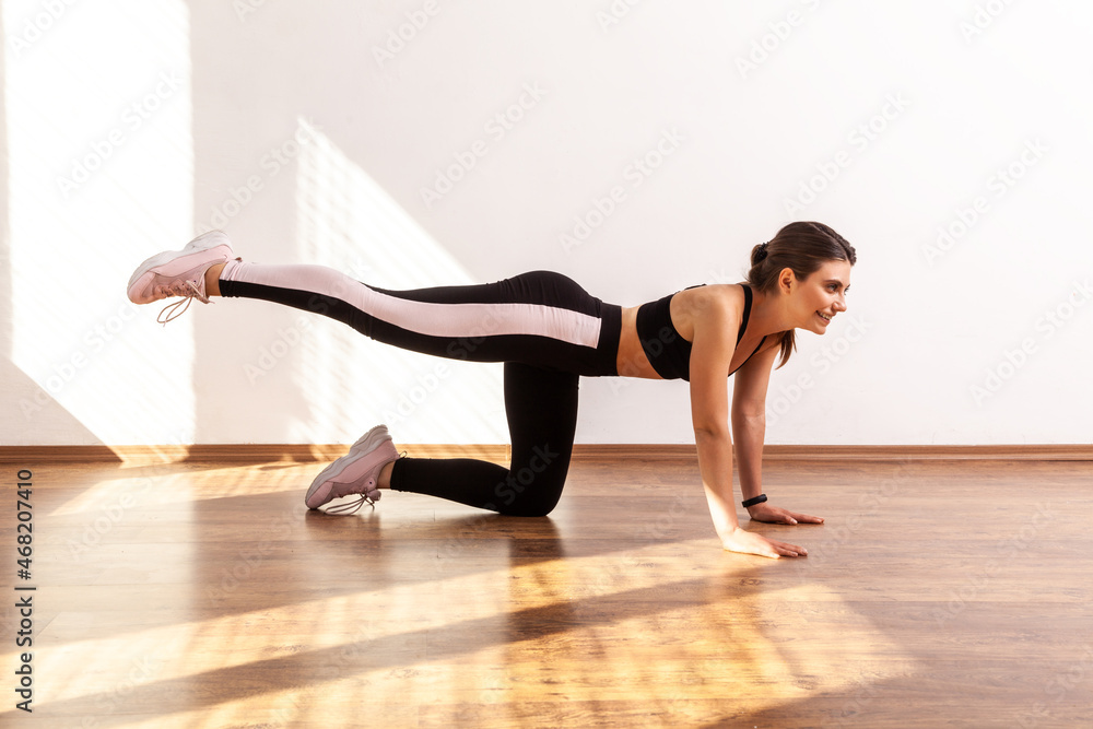 Fit woman practicing sport exercises, doing buttocks and abs workouts at home or fitness gym, wearing black sports top and tights. Full length studio shot illuminated by sunlight from window.