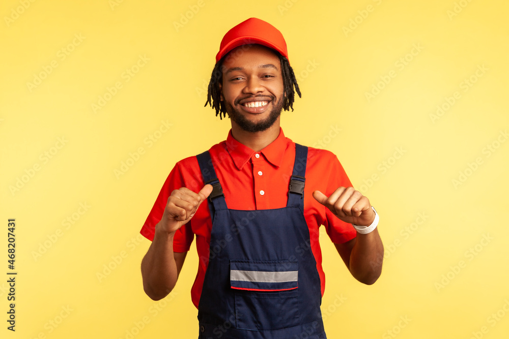 Smiling happy bearded courier or builder wearing overalls, looking at camera with proud expression, pointing at himself, satisfied of work done. Indoor studio shot isolated on yellow background.