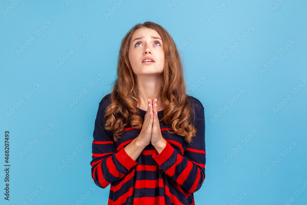 Portrait of young attractive woman wearing striped casual style sweater looking up with imploring desperate grimace, praying to god asking for help. Indoor studio shot isolated on blue background. Portrait of young attractive woman wearing striped casual style sweater looking up with imploring desperate grimace, praying to god asking for help. Indoor studio shot isolated on blue background.