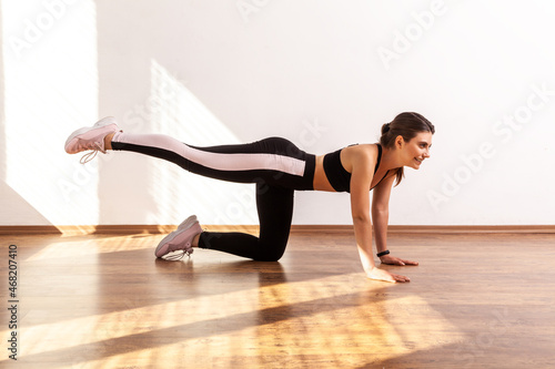 Fit woman practicing sport exercises, doing buttocks and abs workouts at home or fitness gym, wearing black sports top and tights. Full length studio shot illuminated by sunlight from window.