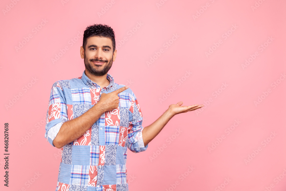 Good looking joyful man with beard in blue shirt presenting advertising area on her palm and pointing to copy space, holding empty place for commercial. Indoor studio shot isolated on pink background. Good looking joyful man with beard in blue shirt presenting advertising area on her palm and pointing to copy space, holding empty place for commercial. Indoor studio shot isolated on pink background.