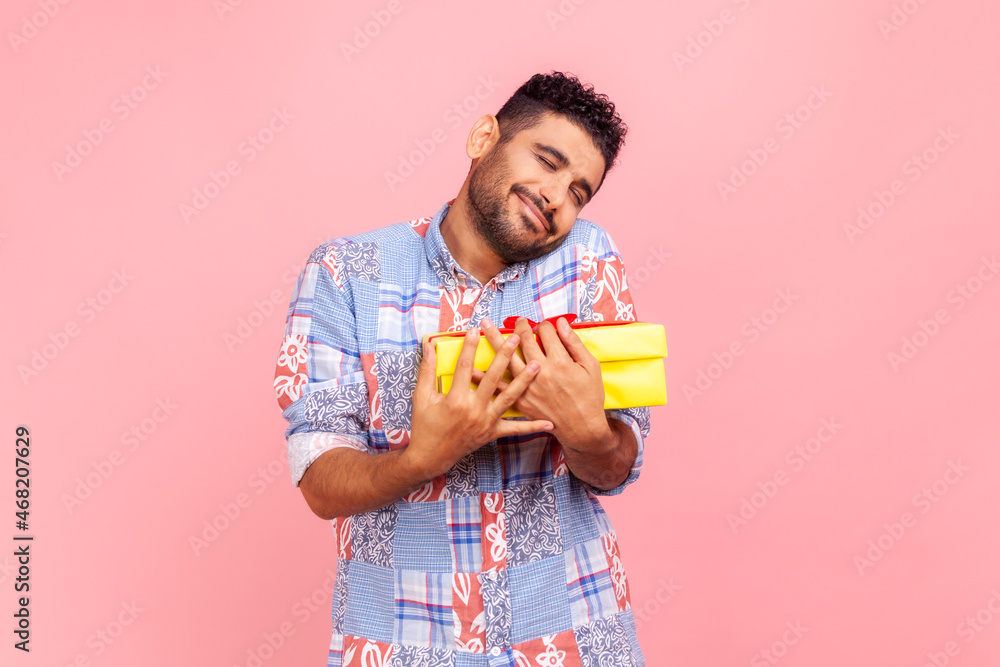 Young adult handsome happy man with beard and dark hair holding embracing yellow wrapped present box, being satisfied to get gift, wearing blue shirt. Indoor studio shot isolated on pink background. Young adult handsome happy man with beard and dark hair holding embracing yellow wrapped present box, being satisfied to get gift, wearing blue shirt. Indoor studio shot isolated on pink background.