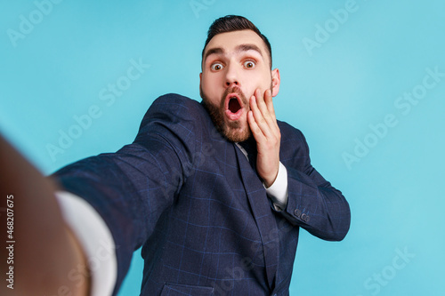Billede på lærred Man with beard wearing dark suit taking selfie or making video call, looking at camera with shocked facial expression POV, point of view of photo, Indoor studio shot isolated on blue background