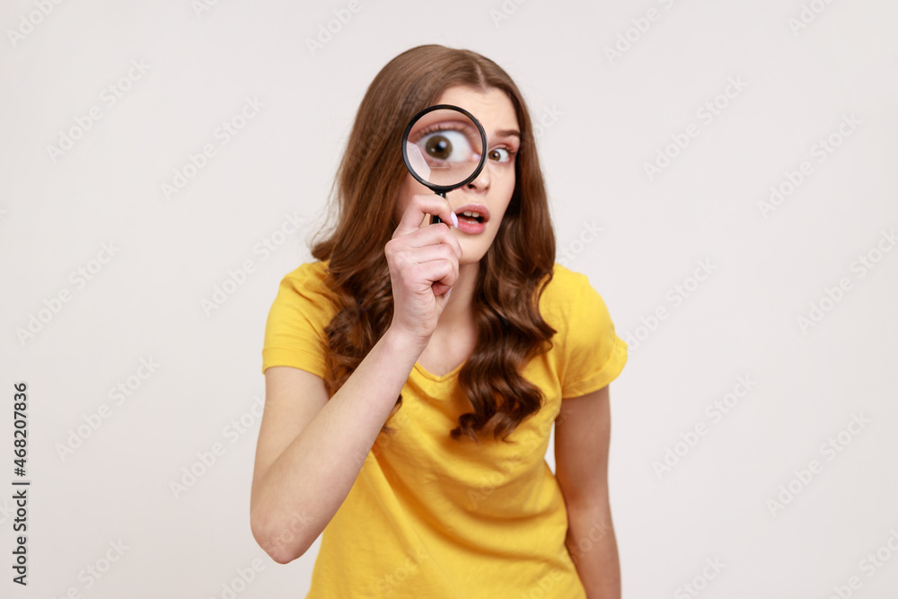 Portrait of funny young female in yellow casual T-shirt holding magnifying glass and looking at camera with big zoom eye, curious face. Indoor studio shot isolated on gray background. Portrait of funny young female in yellow casual T-shirt holding magnifying glass and looking at camera with big zoom eye, curious face. Indoor studio shot isolated on gray background.