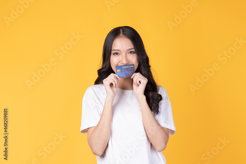 Portrait of a happy young asian woman wearing white shirt holding bank card, credit card isolated on yellow background. Business online shopping concept.