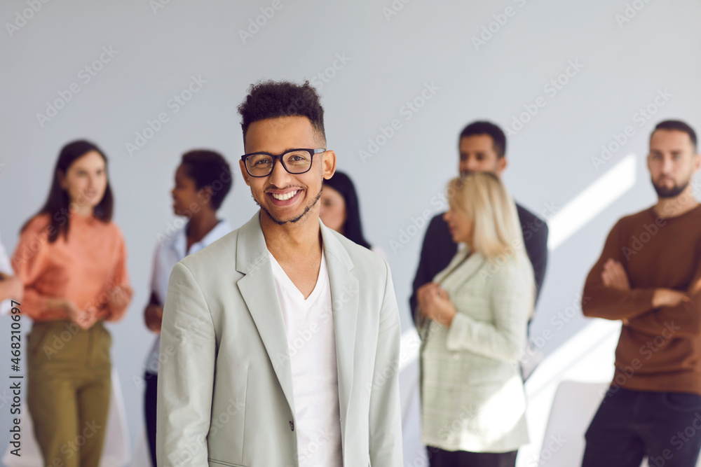 Portrait of positive male employee standing on blurred background of his colleagues. Friendly handsome dark-skinned office worker or businessman working in modern business center. Success concept. Portrait of positive male employee standing on blurred background of his colleagues. Friendly handsome dark-skinned office worker or businessman working in modern business center. Success concept.