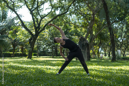 Sporty active young man in black sportswear doing sport exercises in the park. Portrait of handsome fit man doing stretching exercise outdoors. 