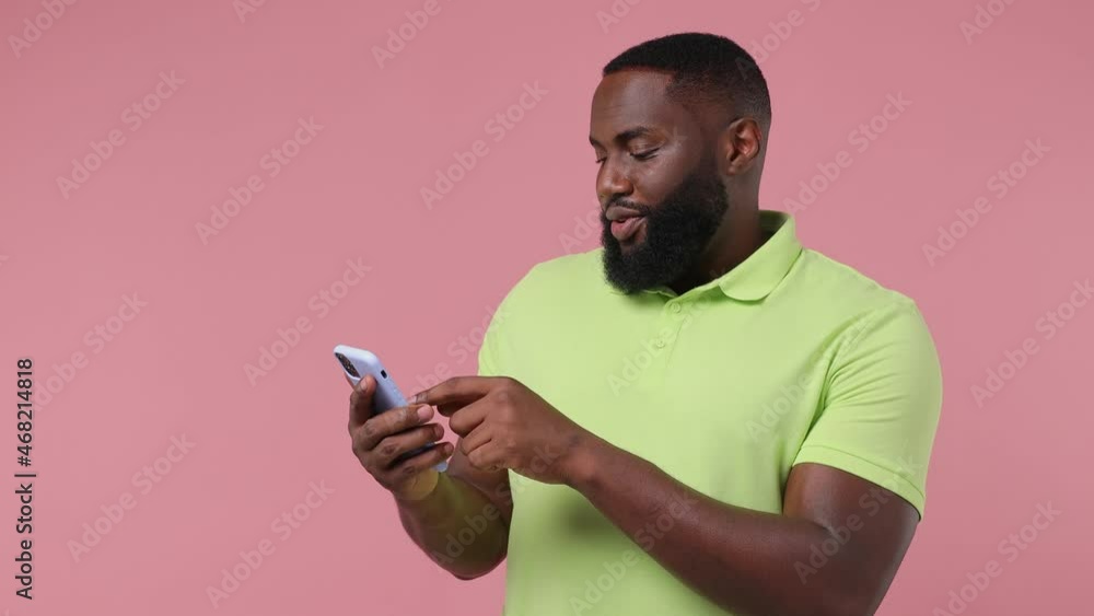 Satisfied young bearded african american man 20s wears green t-shirt hold use mobile cell phone typing browsing swipe do online shopping isolated on plain pastel light pink background studio portrait Satisfied young bearded african american man 20s wears green t-shirt hold use mobile cell phone typing browsing swipe do online shopping isolated on plain pastel light pink background studio portrait