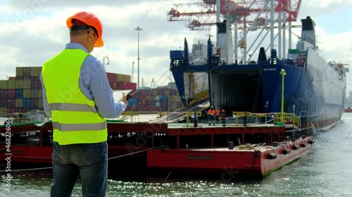 Marine port cargo industry. Safety manager holds tablet computer and portable radio while controls workers team near open ship hold in dock