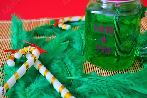 Green drink in a jug decorated with Christmas ornaments and a striped straw surrounded by green feathers and candy canes on a red background