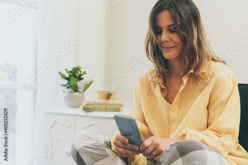 Relaxed girl using smartphone sitting on the sofa at home. Woman chatting with friends sitting on a couch in the living room with warm pajama in a winter morning. Leisure, carefree and relax concept.