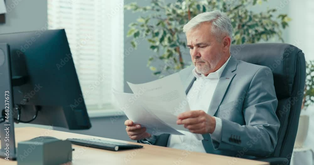 An experienced businessman, grey-haired man head of company is sitting at desk in a comfortable chair of the president, looking at documents brought to him, reading them carefully, asking a colleague An experienced businessman, grey-haired man head of company is sitting at desk in a comfortable chair of the president, looking at documents brought to him, reading them carefully, asking a colleague