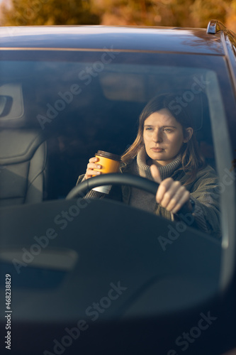 Concentrated young girl in the morning goes to work by car and drinking coffee. View through the windshield. Copy space. Driving car concept.