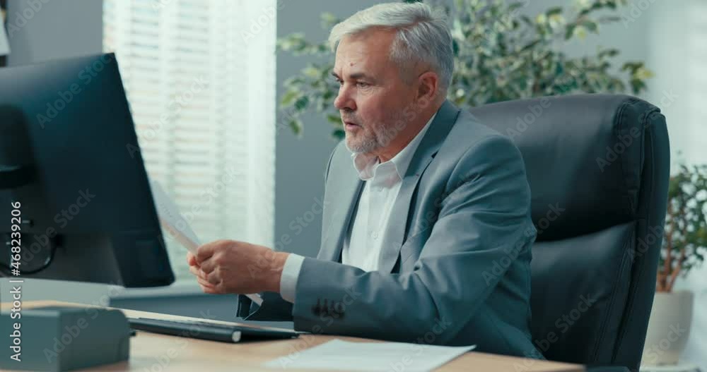 An experienced businessman, grey-haired man head of company is sitting at desk in a comfortable chair of the president, looking at documents brought to him, reading them carefully, asking a colleague An experienced businessman, grey-haired man head of company is sitting at desk in a comfortable chair of the president, looking at documents brought to him, reading them carefully, asking a colleague
