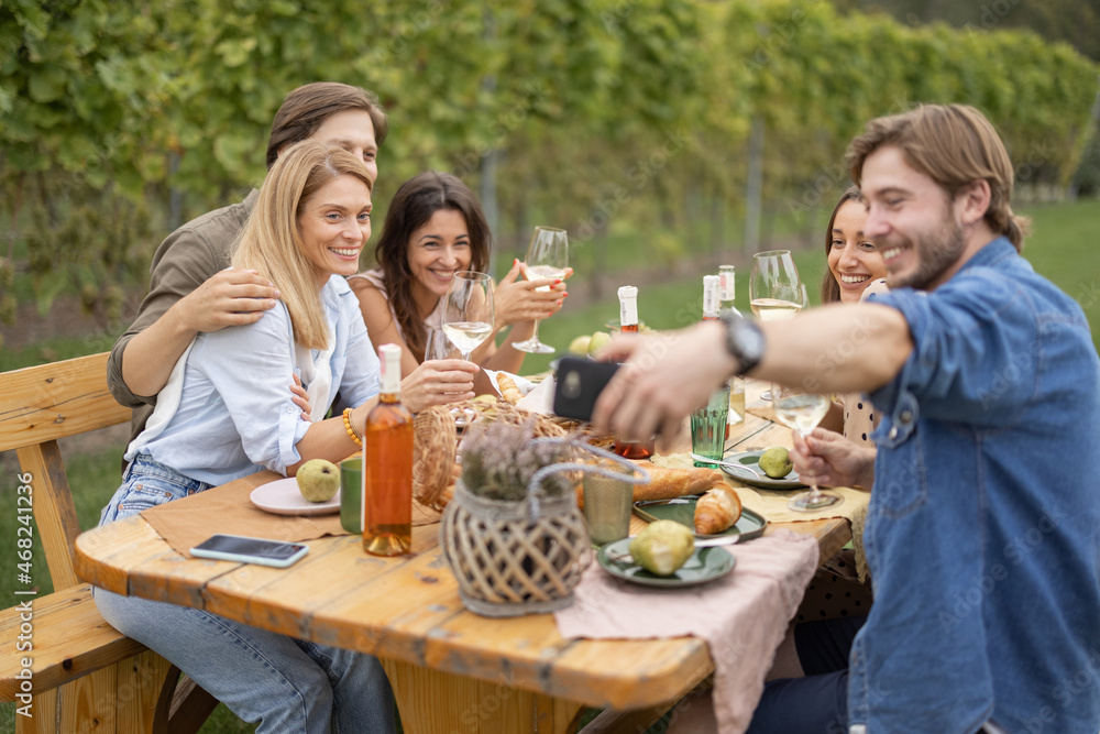 Man taking photo on mobile phone of european friends during friendly picnic. Young men and women drinking local wine from glasses. Concept of friendship. Idea of leisure. Green tourism. Winemaking