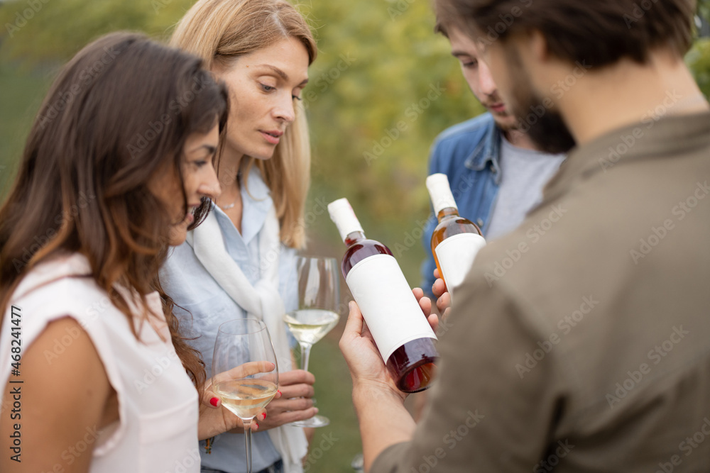 Joyful european friends tasting local wine near vineyards in countryside. Man showing a bottle with blank label to copy paste. Young men and women enjoying time together Joyful european friends tasting local wine near vineyards in countryside. Man showing a bottle with blank label to copy paste. Young men and women enjoying time together