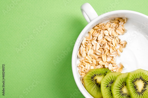 yogurt with kiwi and oatmeal in a white bowl and with oatmeal grains on a green background