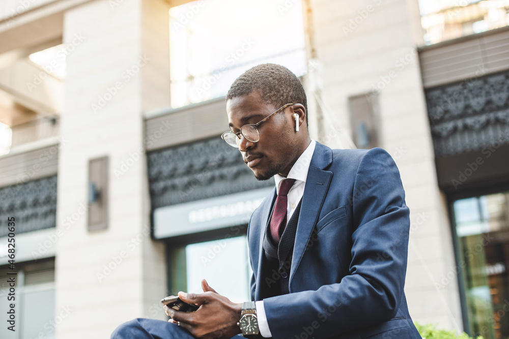 Portrait of black african american businessman in suit with glasses and headphones sits on the background of city buildings outdoors Portrait of black african american businessman in suit with glasses and headphones sits on the background of city buildings outdoors