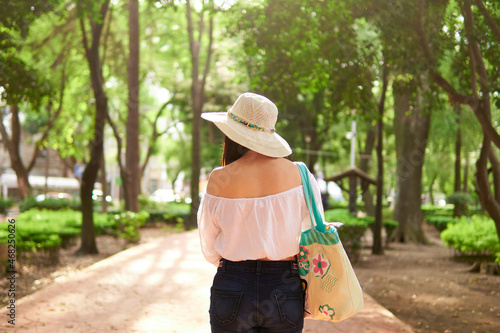 young latin tourist woman walking through a park full of trees at sunset, wearing a hat, blue pants, white blouse and a handbag, model not recognizable, with her back turned, horizontal picture