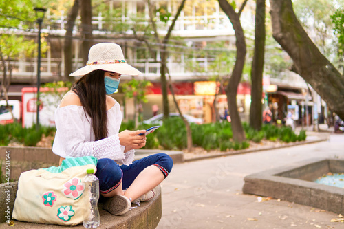 young female tourist sitting in the park with mask sending a message, wearing a hat, blue pants, white blouse, background out of focus, copy space
