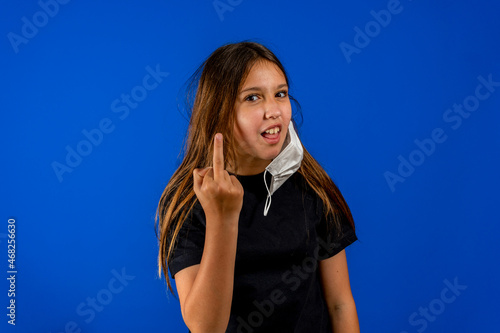 Pretty hispanic preteen girlwearing a half wearing surgical mask to protect herself from the coronavirus doing a fuck you at the camera while smiling, isolated on blue studio background