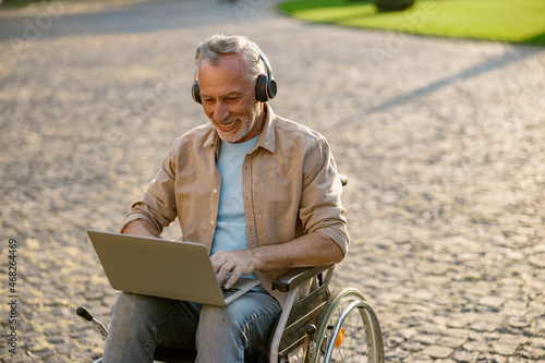 Happy mature man, recovering patient in wheelchair wearing headphones working on a laptop outdoors in the park near clinic