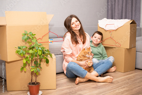 A young beautiful mother with her son moved to a new apartment and are sitting against the background of boxes with their fat ginger cat. photo