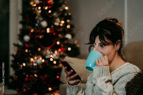 Woman alone at home drinking tea and greeting her family through her smartphone. Christmas time. Candid lights photo