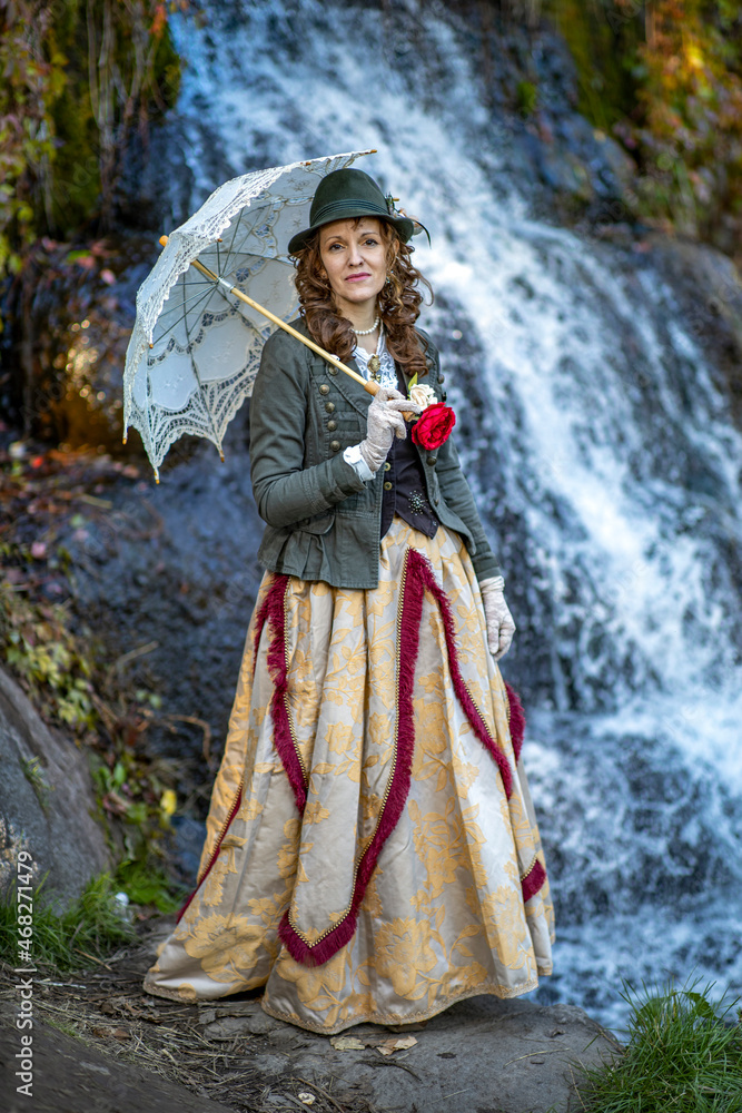 Woman in a 19th century suit with an umbrella against the background of a waterfall in autumn Woman in a 19th century suit with an umbrella against the background of a waterfall in autumn