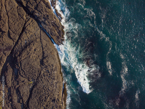 Aerial view. Raging ocean, deep blue water and white foam waves. Rocky dark brown shore. Abstraction. Minimalism. Ecology, environmental protection, beauty of nature. There are no people in the photo