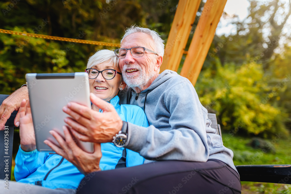 Smiling senior active couple sitting on the bench in the park looking at tablet computer. Using modern technology by elderly. Smiling senior active couple sitting on the bench in the park looking at tablet computer. Using modern technology by elderly.