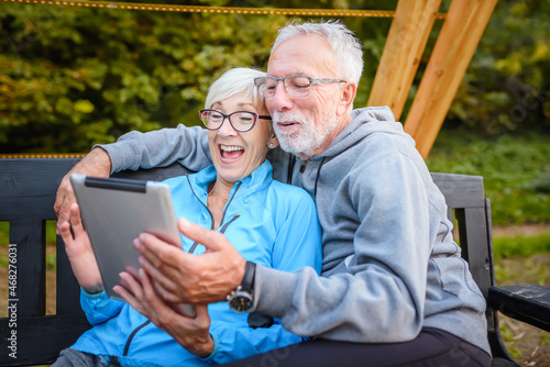 Smiling senior active couple sitting on the bench in the park looking at tablet computer. Using modern technology by elderly.