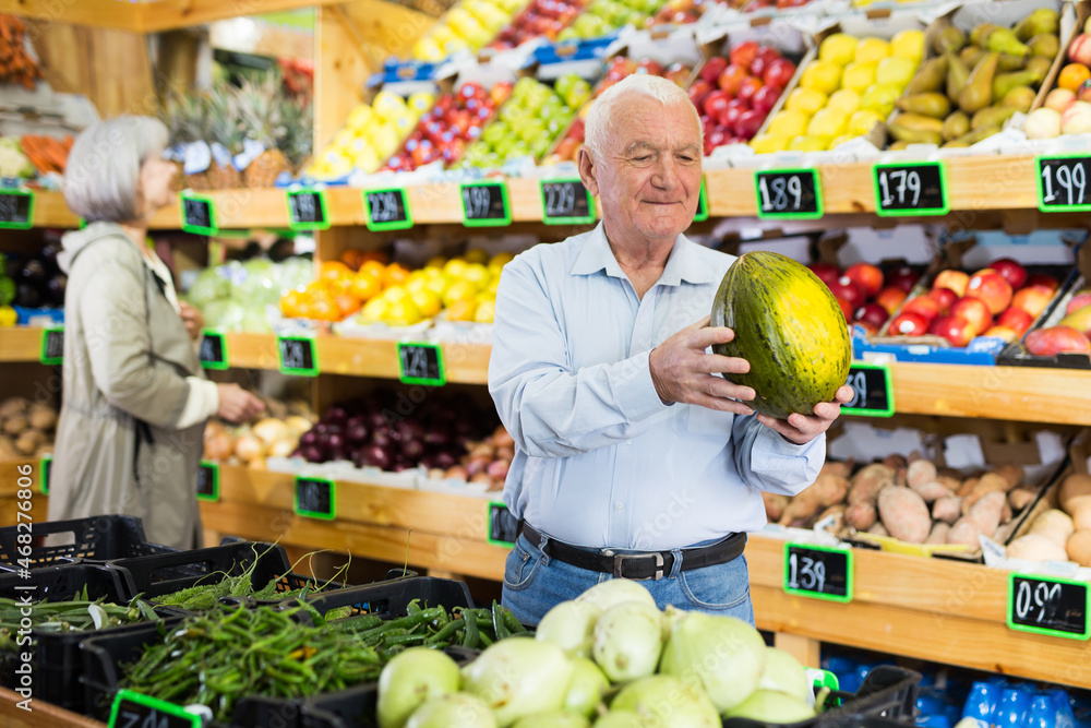 European senior man standing in salesroom of greengrocer with melon in hands. Woman choosing fruits and vegetables in background.
