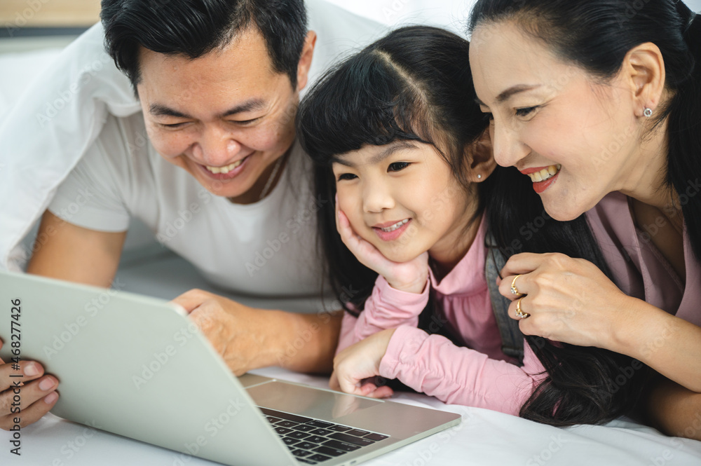 Cheerful young Asian family of handsome father and beautiful mother lying on bed with cute little daughter laughing while watching a comedy movie on laptop and relaxing at home Cheerful young Asian family of handsome father and beautiful mother lying on bed with cute little daughter laughing while watching a comedy movie on laptop and relaxing at home