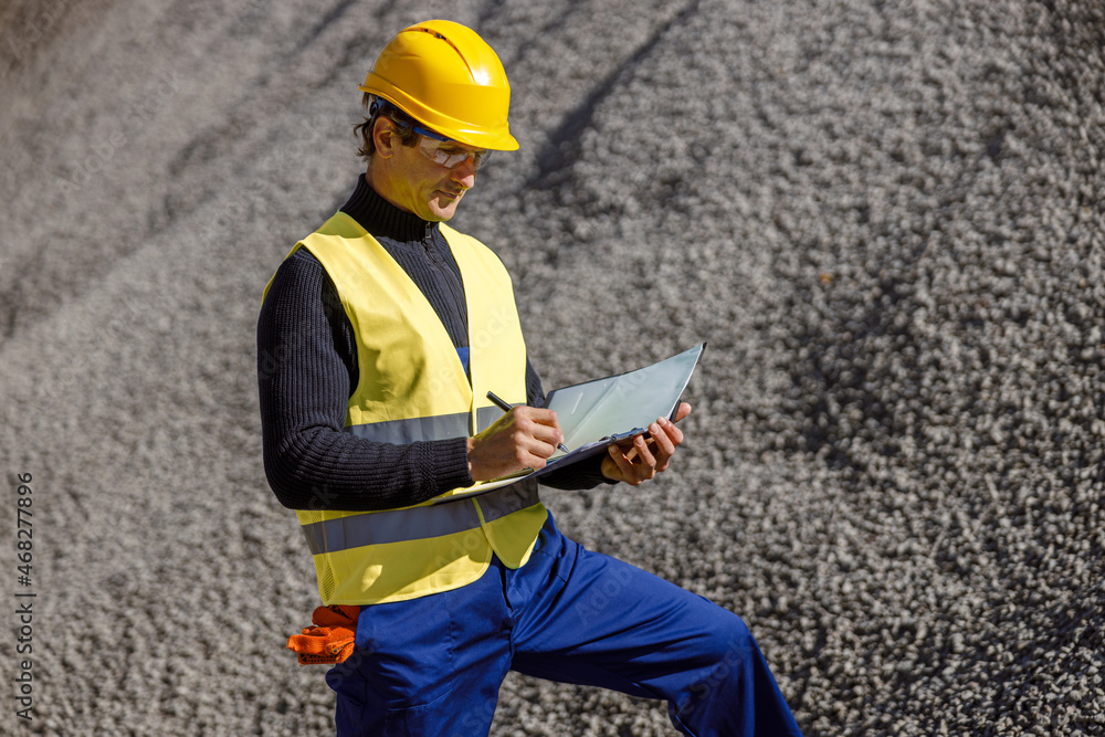 Fototapeta premium Matured man engineer holding folder with papers and taking notes while standing near sand cement mix at concrete plant