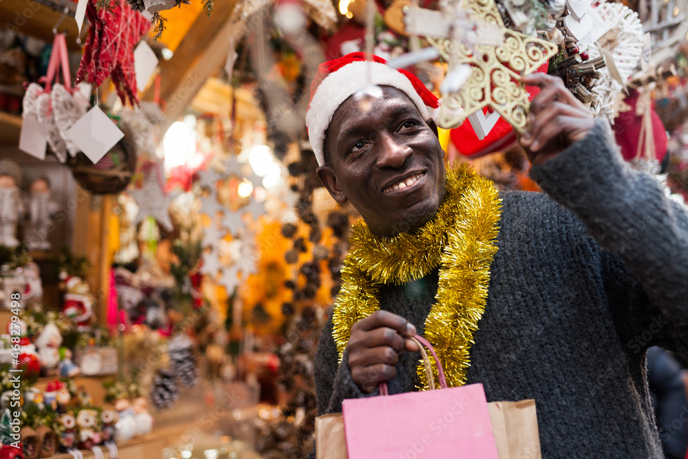 Smiling African American man in Santa hat selecting festive home decoration at outdoor Christmas fair..