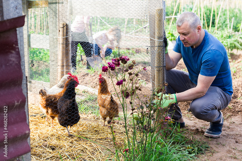 Portrait of positive amateur gardener squatting near chicken coop in his homestead photo