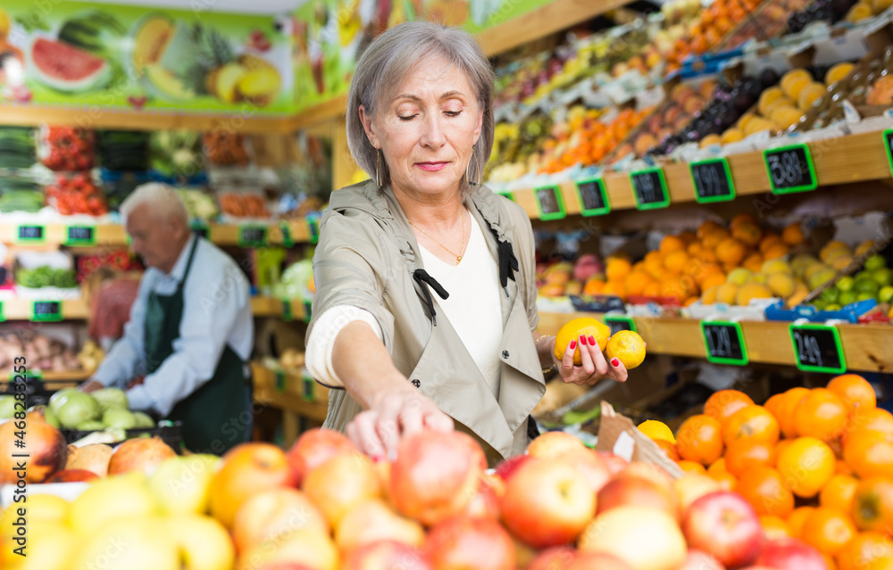 Old lady choosing fruits while standing in salesroom of greengrocer. Senior merhcandiser working in background.