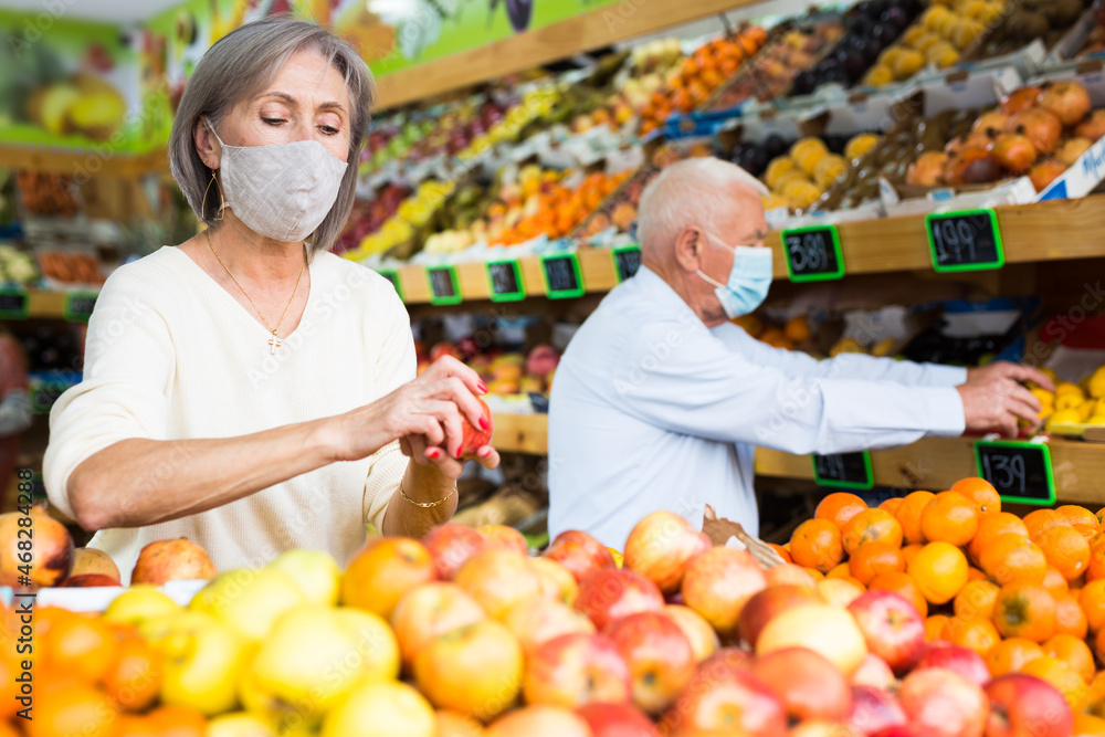 Adult woman in protective mask choosing sweet apples and other fruit on counter of farmers market