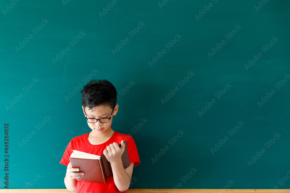 Asian boy holding book standing in front of green board in summer spacial classroom, back to school concept .
