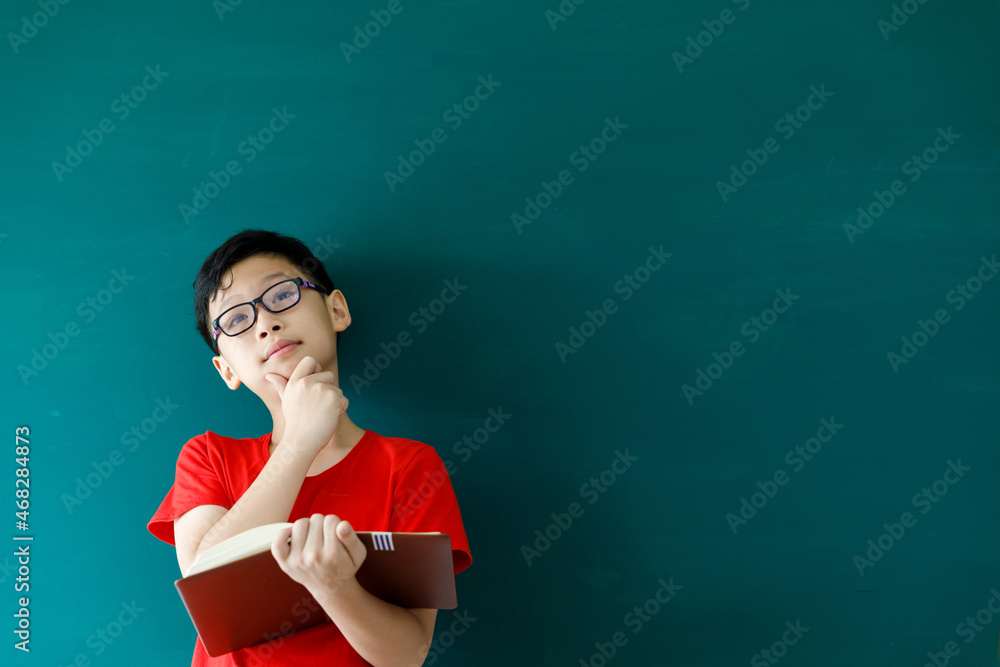 Asian boy holding book standing in front of green board in summer spacial classroom, back to school concept .