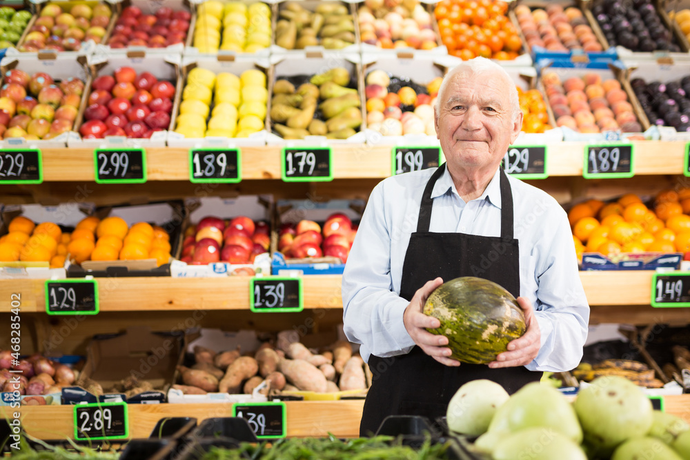 European senior man supermarket worker standing in salesroom and holding melon in hands.