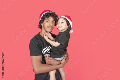 immigrant family of brown latino boy with christmas hat holding smiling child, pink background with copy space © SETO fotografias